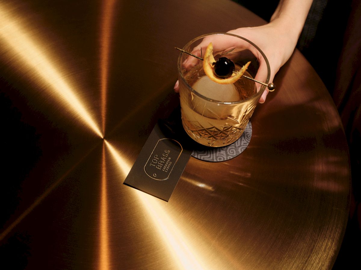 A hand holds a decorative glass cup with a curled rim and a black tag on a shiny, brushed metal surface. Top it at 140 characters, always ending the sentence.