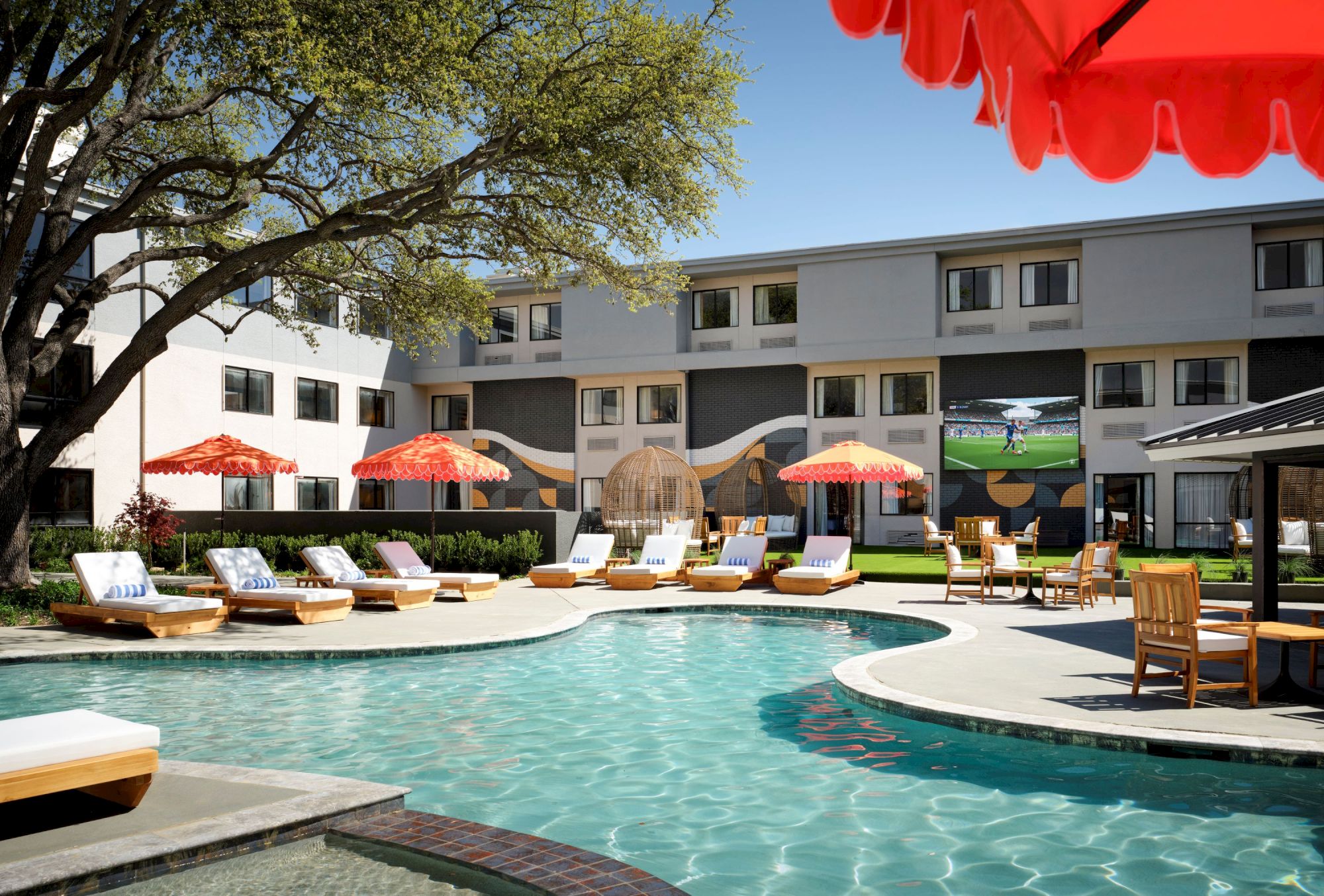 A sunny motel-style pool area with lounge chairs, orange umbrellas, a curved pool, and a two-story white building in the background, under a tree.