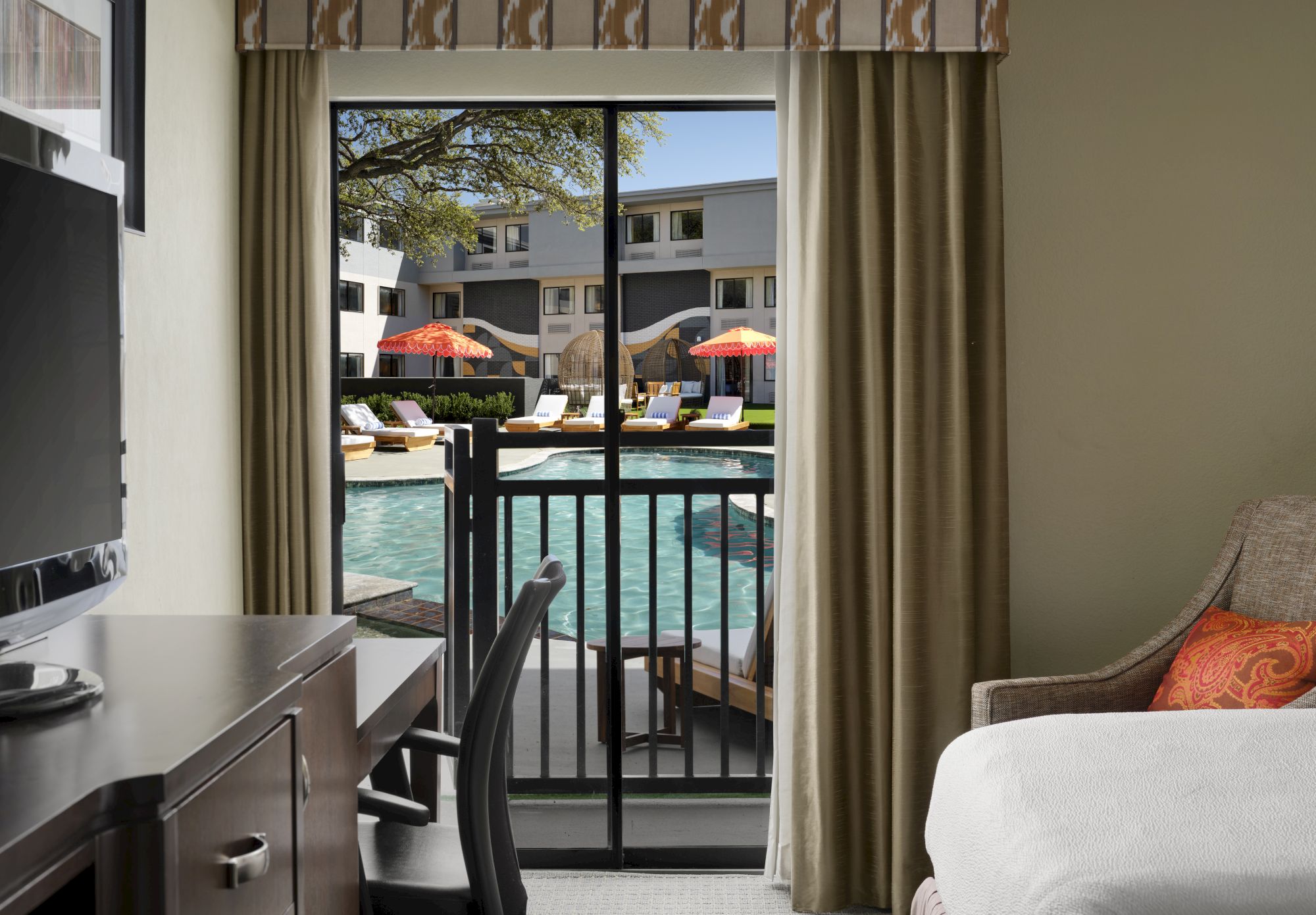 A hotel room with a sliding glass door opening to a balcony overlooking a pool area with lounge chairs and umbrellas.
