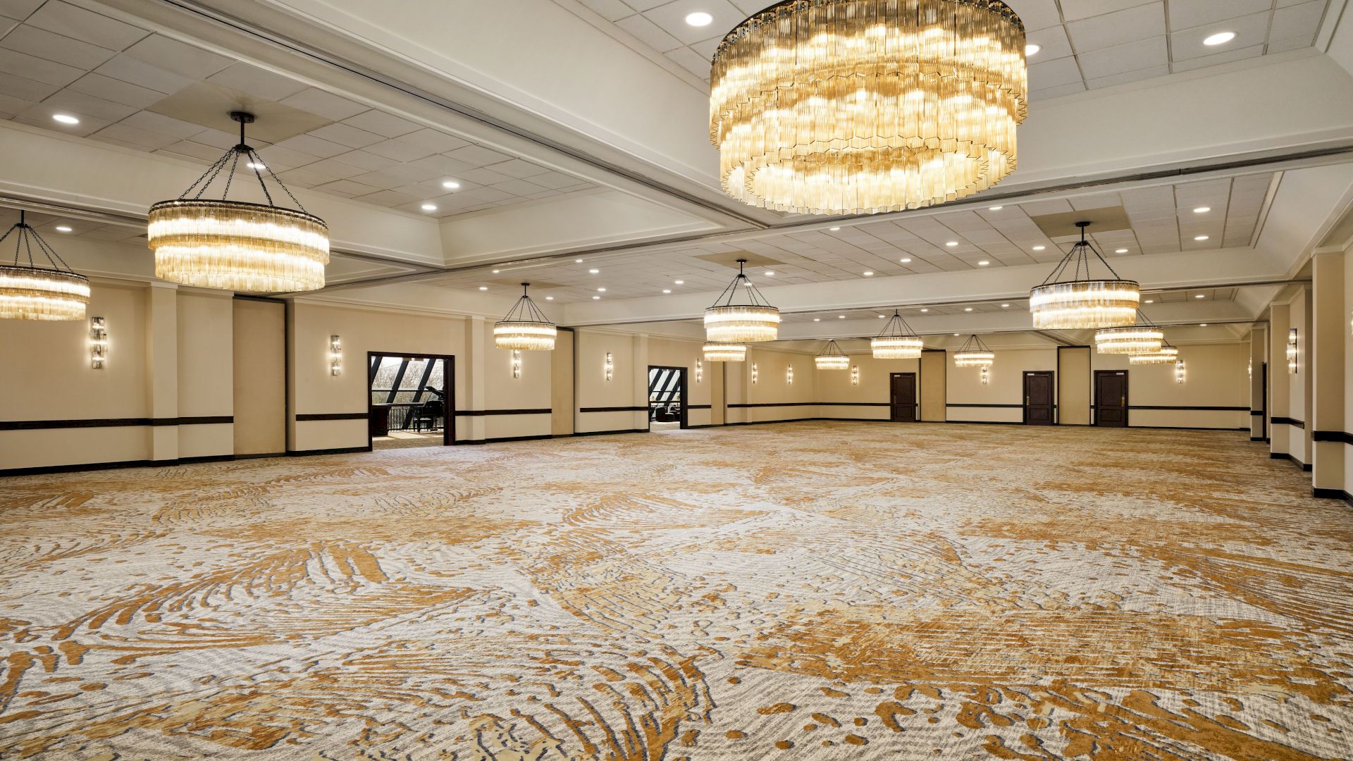 A grand hotel ballroom with ornate gold beaded chandeliers hanging above patterned carpeted floors.