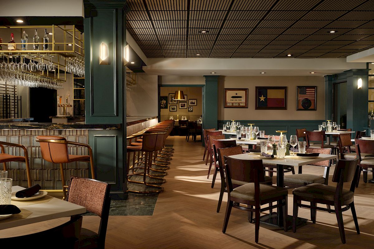 A stylish, dimly lit restaurant with green-paneled walls, wooden tables set with white napkins, and a bar area on the left; cozy, elegant dining ambiance.