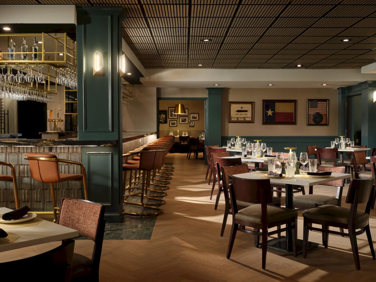 A stylish, dimly lit restaurant with green-paneled walls, wooden tables set with white napkins, and a bar area on the left; cozy, elegant dining ambiance.