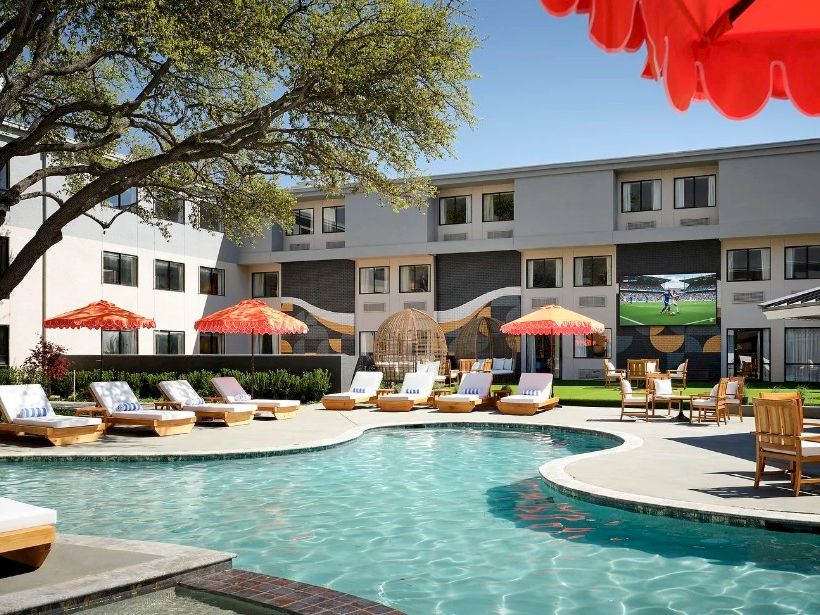 A hotel pool area with lounge chairs, orange umbrellas, and a multi-story motel-style building in the background, under a sunny sky.
