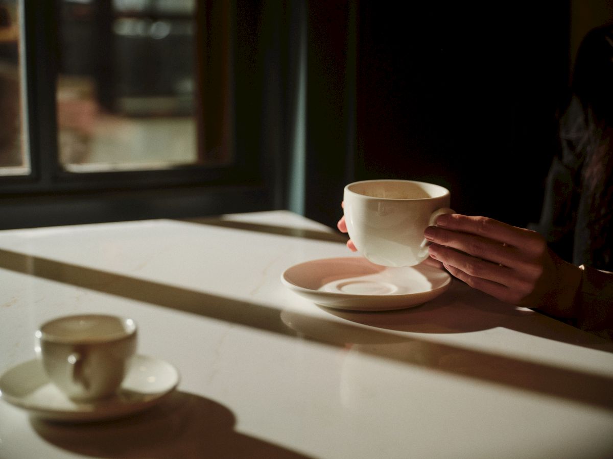 Two people are sharing a quiet moment with white teacups on a sunlit table near a window, hands cradling a cup on a saucer.
