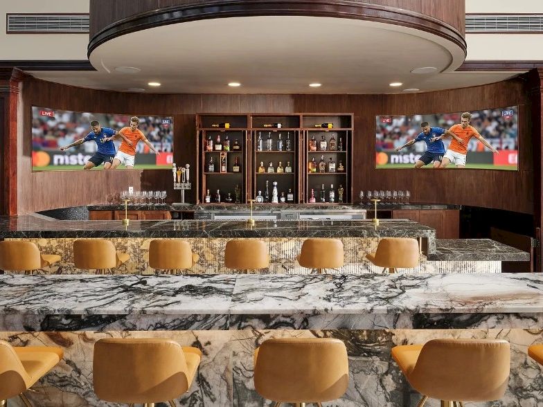 A sleek marble bar with tan stools, backlit shelves, two TVs showing sports, and a polished, upscale lounge vibe.