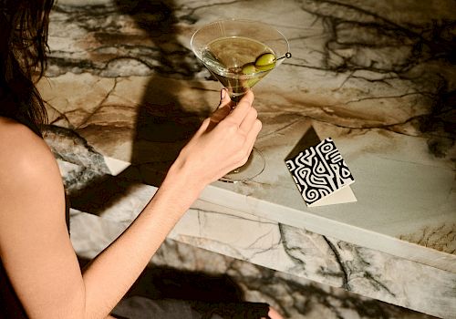 A person holds a martini glass over a marble surface with a black and white patterned card nearby.