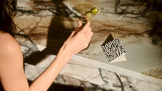 A person holds a martini glass over a marble surface with a black and white patterned card nearby.