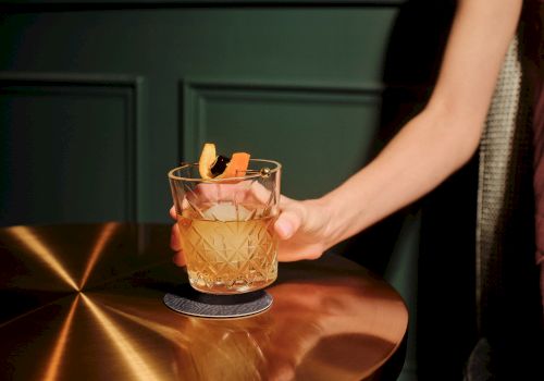 A neatly carved whiskey on a copper bar, garnished with an orange twist, hand resting nearby, cozy bar vibe.