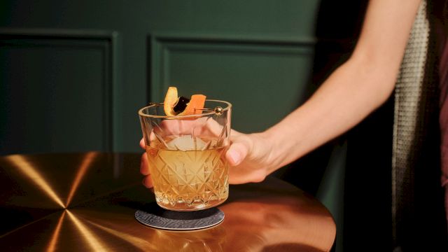 A neatly carved whiskey on a copper bar, garnished with an orange twist, hand resting nearby, cozy bar vibe.