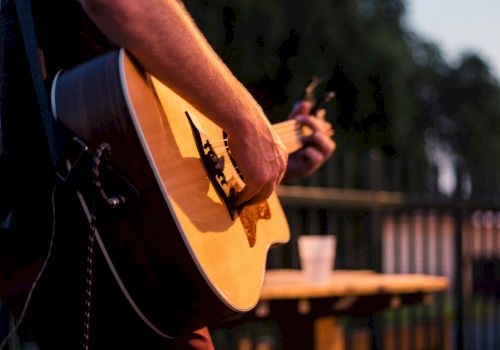 A close-up of a person playing an acoustic guitar outdoors, focus on the strings and body, with a blurred background and a table in view, craft note.