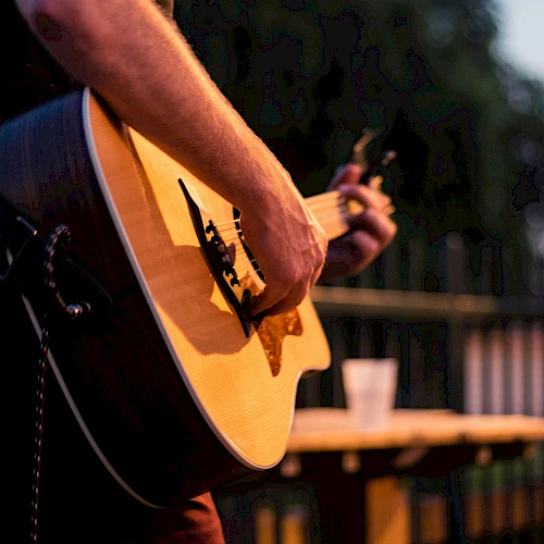 A close-up of a person playing an acoustic guitar outdoors, focus on the strings and body, with a blurred background and a table in view, craft note.