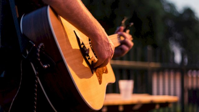 A close-up of a person playing an acoustic guitar outdoors, focus on the strings and body, with a blurred background and a table in view, craft note.