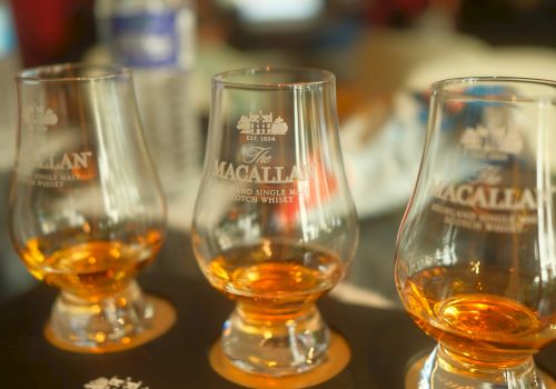 Three whisky glasses with amber liquid on a Matagallan bar tray, close-up, warm bokeh background, tasting setup, Macallan branding.