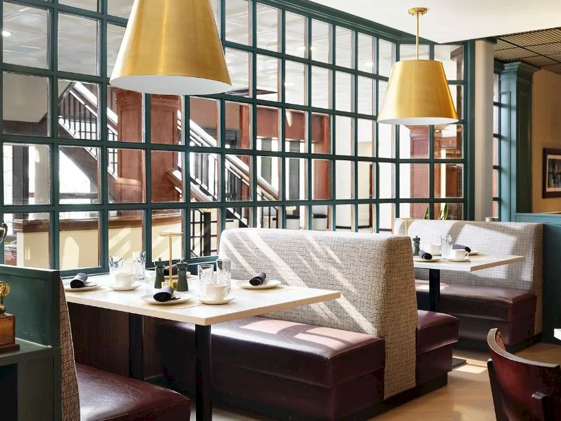 Cozy restaurant booth seating with cream upholstery, large grid windows, and warm pendant lights overhead, creating a bright, inviting dining space.