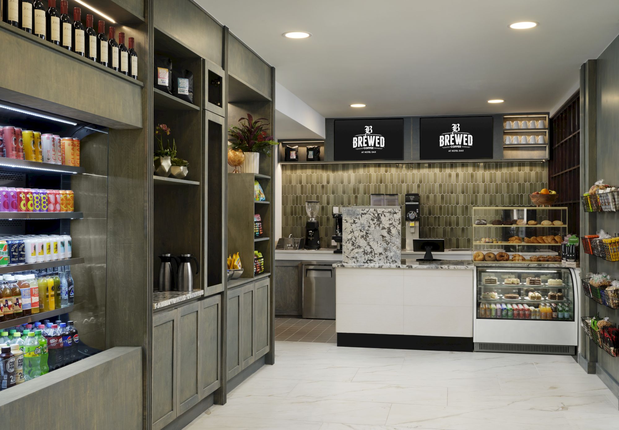 A modern cafe or bakery shop with a clean, gray-toned interior, a central counter, pastry case, shelves of drinks, and warm lighting.