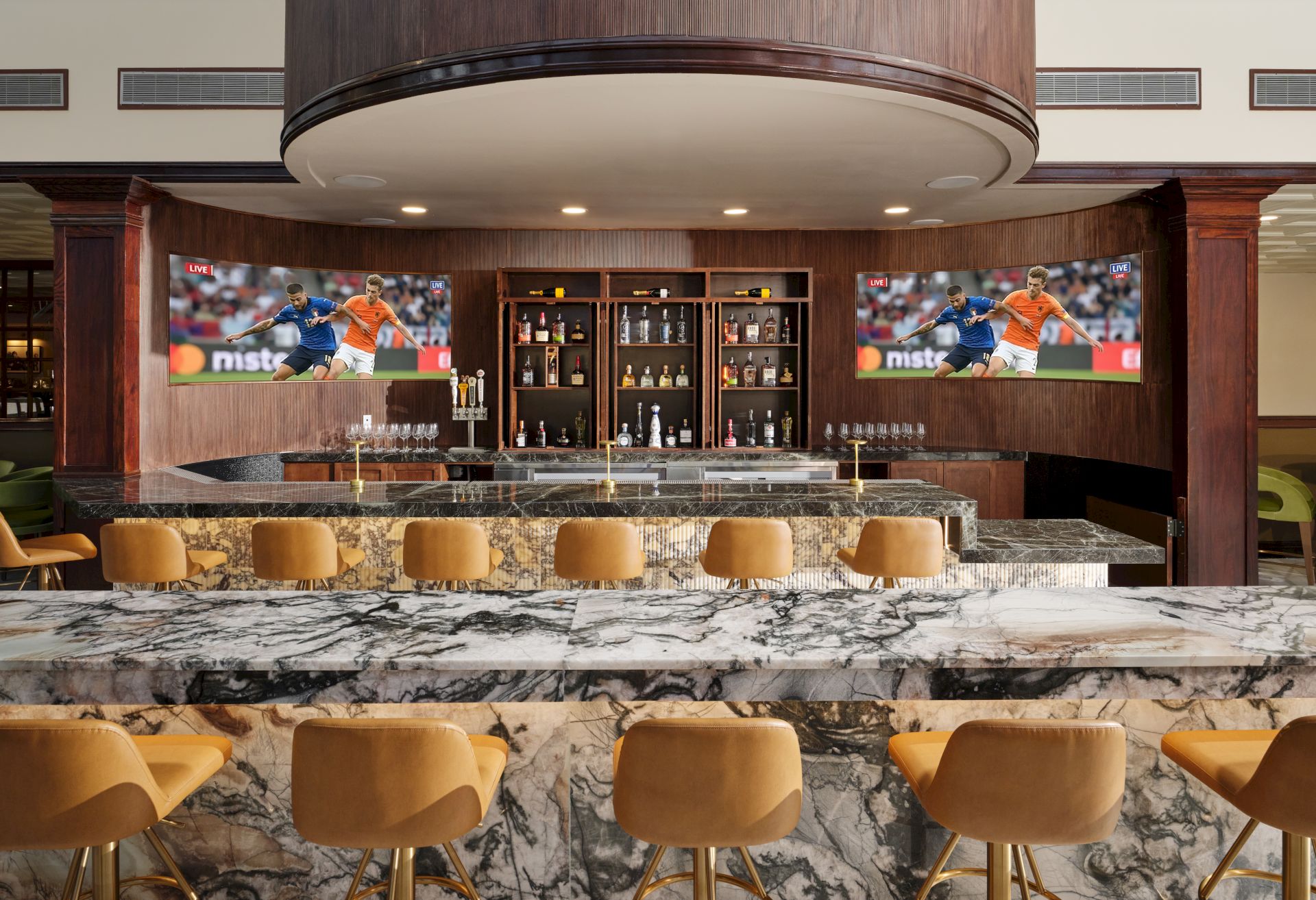 A stylish hotel bar with a marble counter, liquor bottles, and two screens showing a live soccer match, under warm recessed lights.