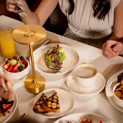 People dining on a table filled with assorted dishes, including toast, fruit, salad, a candleholder, coffee, and drinks, in a cozy meal setting.