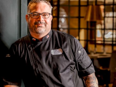 Chef in black uniform with name tag, standing in a kitchen doorway, arm tattoos, warm lighting and wooden decor behind him.