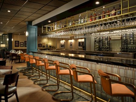 A stylish bar interior with a long tiled counter, orange swivel stools, hanging wine glasses, backlit shelves, and intimate seating in the background.