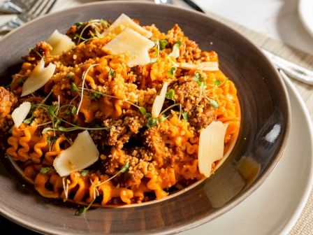 Pasta with meat sauce, grated cheese shavings, and herbs on a dark plate, a hearty Italian-inspired dish served warm.