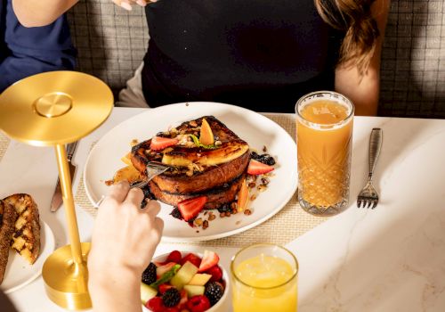 A table with a plated breakfast: toast with fruit and syrup, berries in a bowl, orange juice, and a golden candle holder on a white marble table.