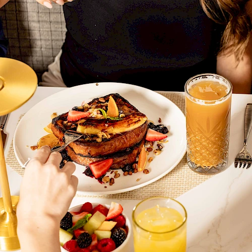 A table with a plated breakfast: toast with fruit and syrup, berries in a bowl, orange juice, and a golden candle holder on a white marble table.