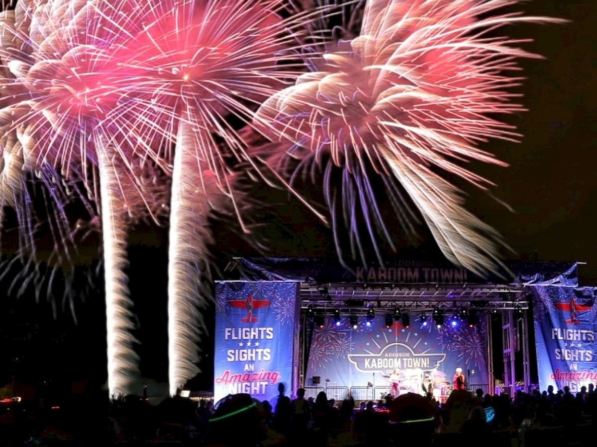 Fireworks explode above a crowd at a night festival or concert stage with blue banners and a lit audience.