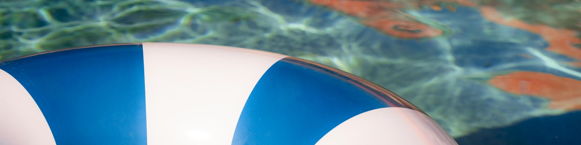 A blue and white striped life buoy floats near the edge of a clear swimming pool, with sunlight reflecting off the blue water.