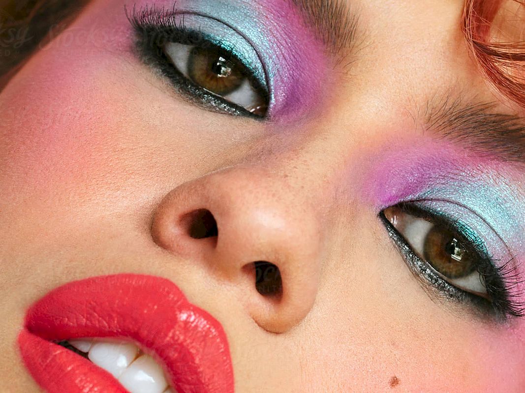 Close-up of a person with bold makeup: pink, purple, and blue eyeshadow, red lips, and dramatic eyeliner.