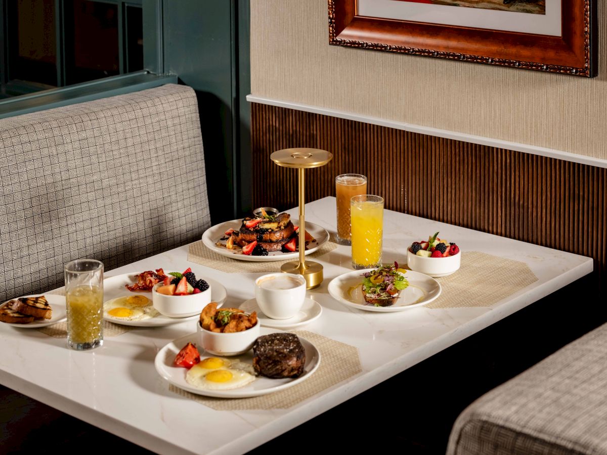 A deluxe brunch spread on a booth table: assorted pastries, fruits, a chocolate dessert, mini burgers, and several plates with juice and coffee, all neatly arranged.