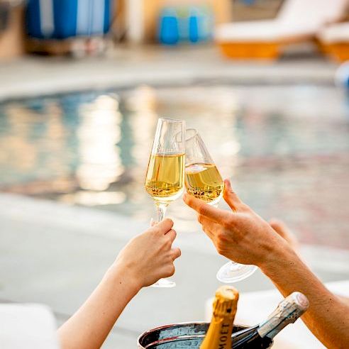 People cheers with champagne glasses by a poolside; a bucket of bottles sits nearby as they celebrate together by the water.