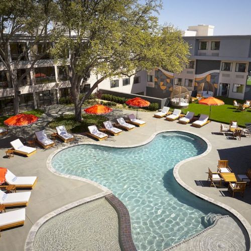 An outdoor hotel pool area with a unique curved shape, sun loungers, orange umbrellas, and a grassy courtyard in a sunny courtyard.