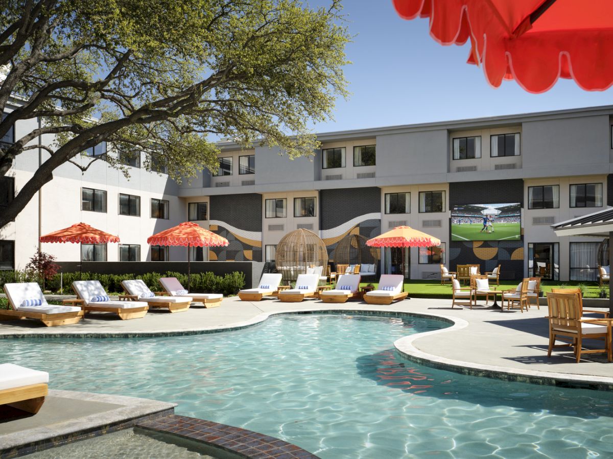 A sunny hotel pool area with lounge chairs, umbrellas, and a two-story building in the background, under a clear sky, sunny day.