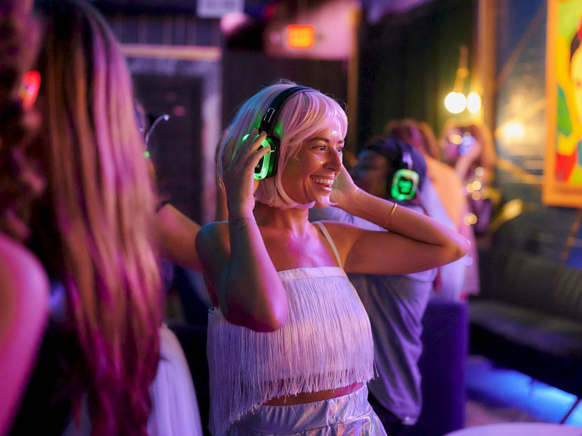 People dancing in a club with neon lights; a smiling woman wearing headphones, enjoying the music at a lively party.