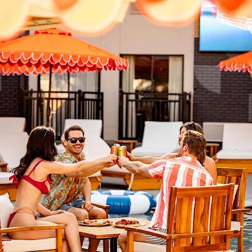 Four friends toast drinks by a poolside under red umbrellas, sitting on wooden chairs with a sunny resort vibe.