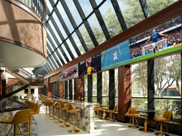 A modern, sunlit sports lounge with yellow chairs along a long glass wall, TV screens showing a tennis match, and a marble bar in a bright, airy setting.