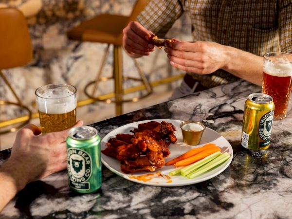 Two people at a marble table share wings, celery sticks, and dips, with beer cans and a glass of beer on the side.