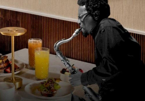 A person wearing dark clothing is playing a saxophone-style instrument at a dining table with orange juice and plates of food, in a hotel-like setting.