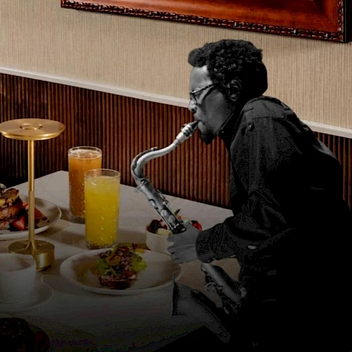 A person wearing dark clothing is playing a saxophone-style instrument at a dining table with orange juice and plates of food, in a hotel-like setting.