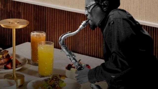 A person wearing dark clothing is playing a saxophone-style instrument at a dining table with orange juice and plates of food, in a hotel-like setting.