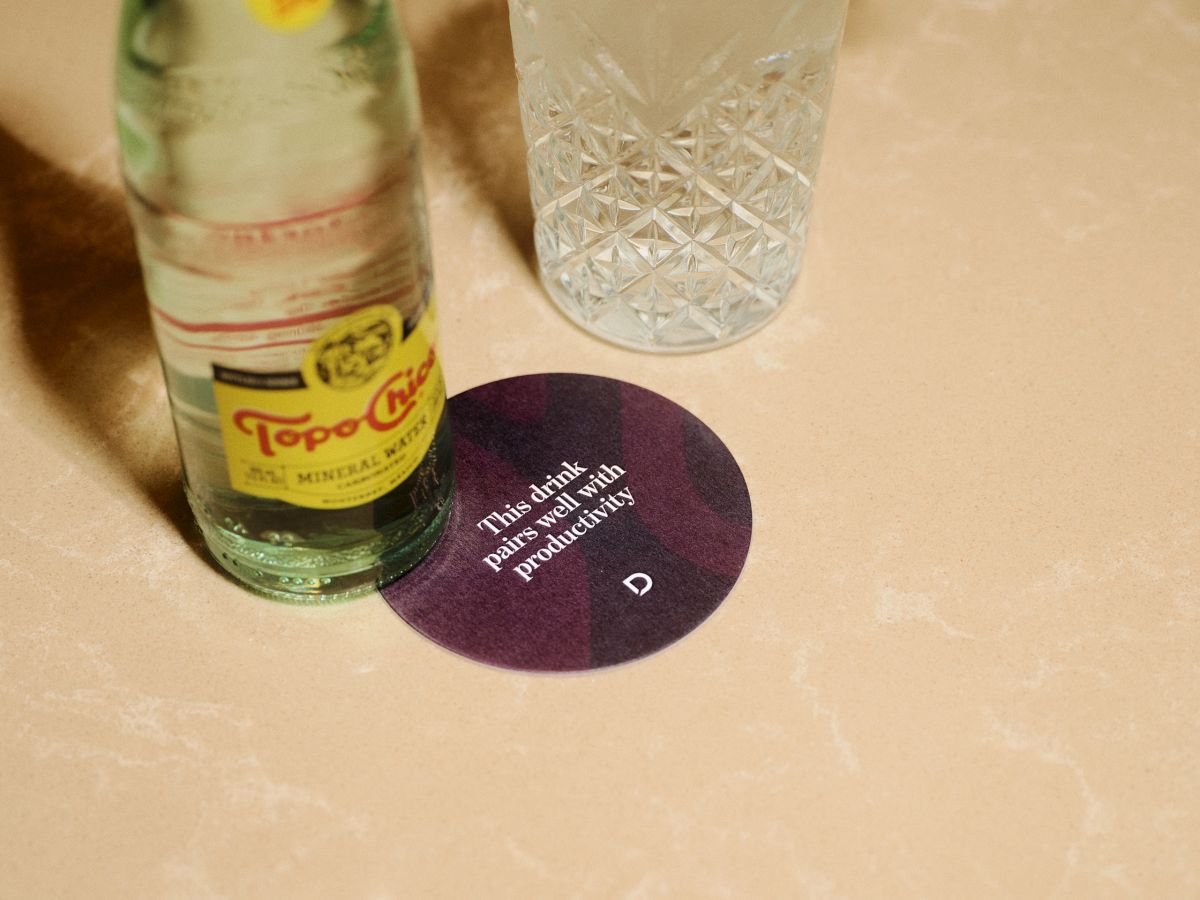 A bottle of Topo Chico-like mineral water beside a cut-glass glass on a table, with a purple circular coaster or card between them.