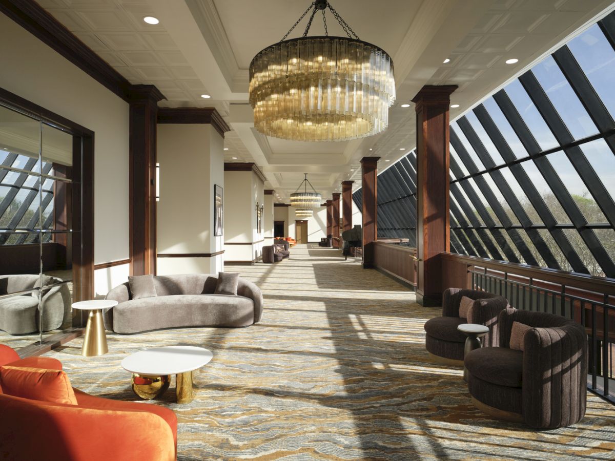 A stylish, sunlit hotel lounge with modern seating, chandeliers, and a glass railing overlooking a slanted window wall. End of sentence.