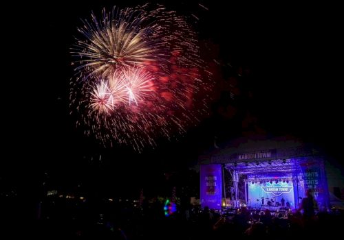 Fireworks explode above a crowd at night, while a lit stage with a blue glow hosts a live outdoor performance. End sentence.