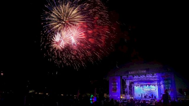 Fireworks explode above a crowd at night, while a lit stage with a blue glow hosts a live outdoor performance. End sentence.