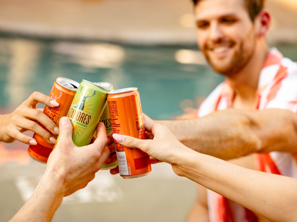 A group of friends cheers with colorful soda cans by a pool, clinking them together as one smiles in a summer celebration.