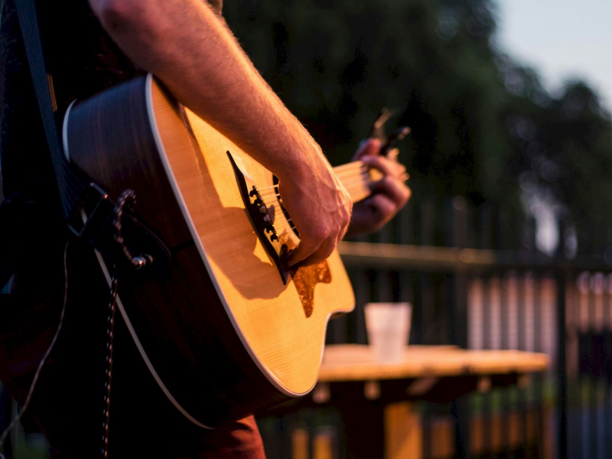 A guitarist strums an acoustic guitar on an outdoor stage, with a blurred background and wooden table nearby.