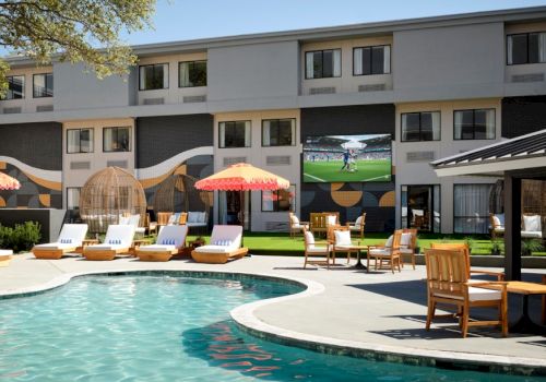 A sunny hotel pool area with lounge chairs, umbrellas, and a European-style courtyard; guests relax while a flat-screen TV shows a sports game.