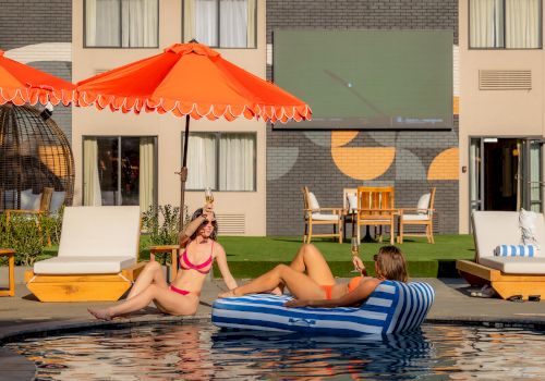Two women lounge poolside on a sunny day under an orange umbrella, sipping champagne in bikinis while others relax on sunbeds.