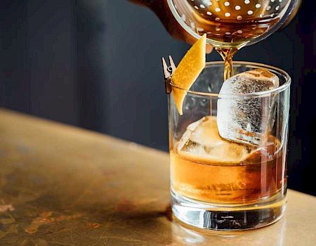 A bartender pours amber cocktail into an ice-filled rocks glass, garnished with a citrus wedge.