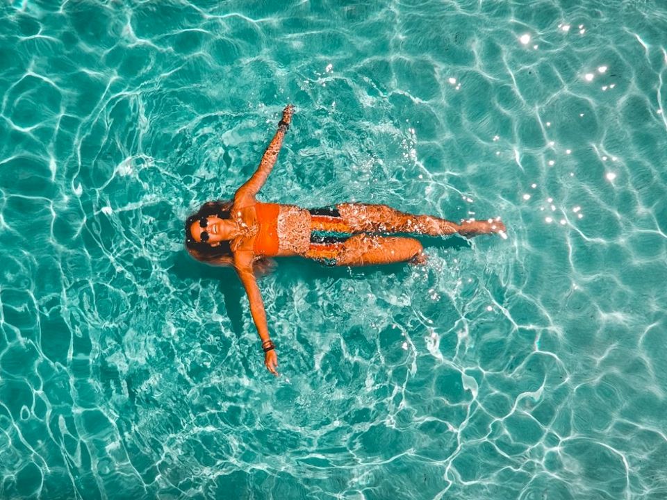 An aerial view shows a person floating face up in clear turquoise ocean water, surrounded by rippling waves.